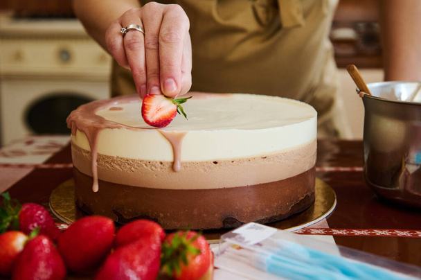 Γκρο πλαν άποψη ενός ζαχαροπλάστη σεφ βάζοντας φρέσκια φέτα φράουλας πάνω από μια τούρτα γενεθλίων, στρωμένη με τρεις στρώσεις μους σοκολάτας, στην κουζίνα come. Ζαχαροπλαστική. Αρτοποιείο. Βιομηχανία τροφίμων - Φωτογραφία, εικόνα
