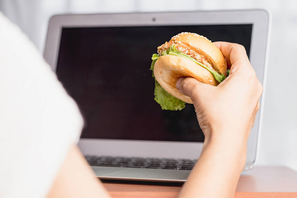 Hand houden van een heerlijke hamburger en het gebruik van laptop computer om te eten tijdens het werk concept. - Foto, afbeelding