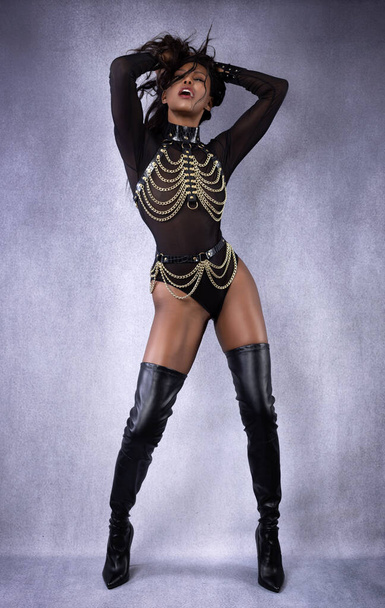 Beautiful sexy black dominant woman in leather chain harness outfit and thigh high boots posing full length in studio on gray mottled background - Photo, image