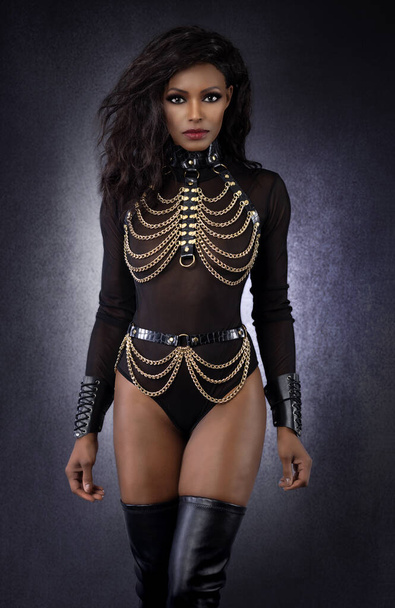 Portrait of beautiful sexy black dominant woman in leather chain harness outfit and thigh high boots posing in studio on gray mottled background - Fotó, kép