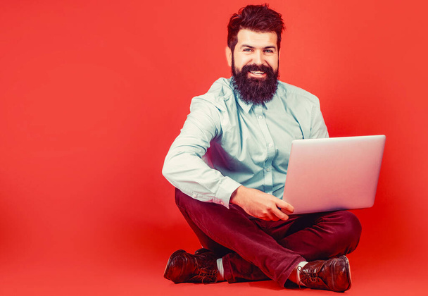 Szczęśliwy młody człowiek siedzący na podłodze i korzystający z laptopa na czerwonym tle. Trzymanie laptopa. - Zdjęcie, obraz
