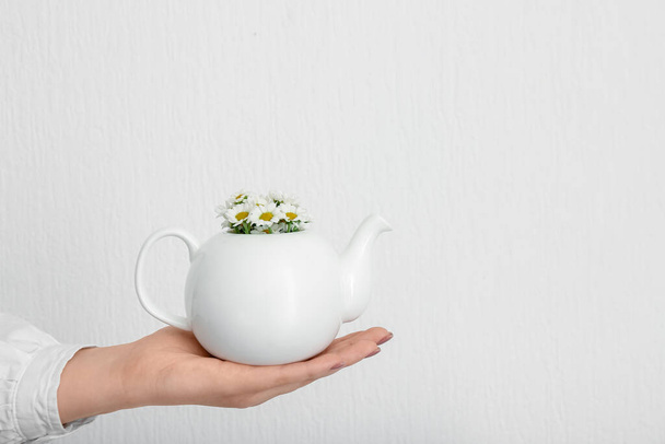 Female hand with teapot of chamomile tea and flowers near white wall - Fotó, kép