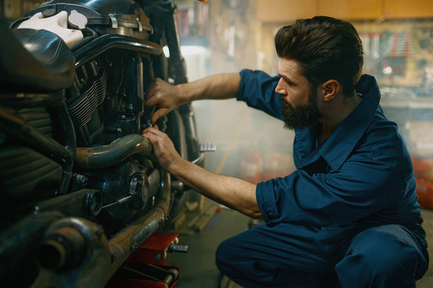 Молодой механик носит комбинезон, используя профессиональные инструменты, работающие на мотоцикле в ремонтной службе гаража - Фото, изображение