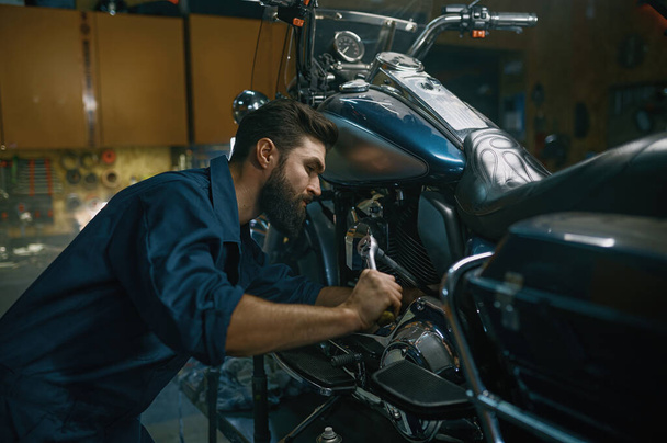Man technician repairing motorcycle using wrench while working at repair workshop. Professional maintenance of modern and vintage motorbike concept - Foto, Imagen