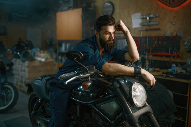 Портрет жестокого небритого механика, сидящего на отремонтированном мотоцикле на фоне мастерской - Фото, изображение