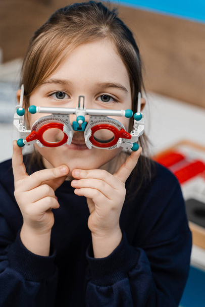 Dítě ve zkušebním rámu brýle na modrém pozadí. Vyšetření s pediatrickým oftalmologem pro výběr zkušebního rámu brýlí pro vyšetření zrakového systému - Fotografie, Obrázek
