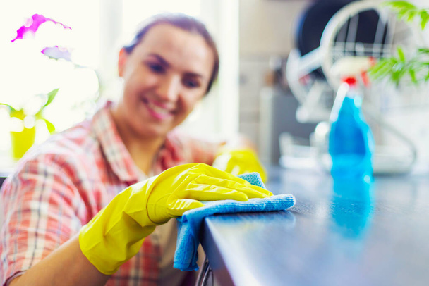 Νοικοκυρά με κίτρινα γάντια σκουπίζει τη σκόνη χρησιμοποιώντας απορρυπαντικό ψεκασμού και κουρέλι. Η γυναίκα κάνει δουλειές του σπιτιού. Έννοια καθαρισμού - Φωτογραφία, εικόνα