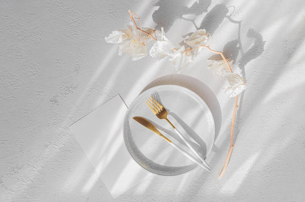 Festive table setting. Golden modern cutlery, dried orchid branch, ceramic plate and blank paper as mock up for invitation or congratulation with beautiful shadows from sunlight on white background - Photo, Image