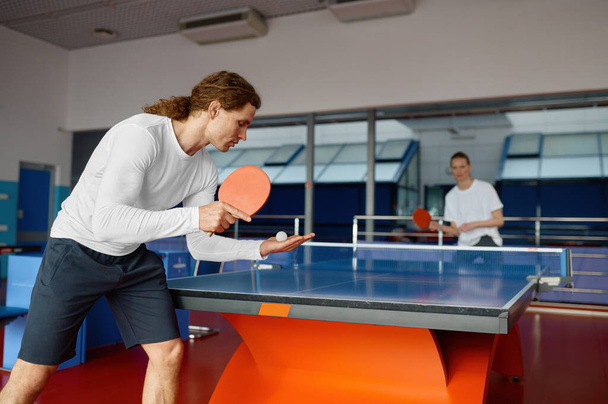 男性と女性はテーブルテニスをプレイし、ラケットでボールを提供するスポーツマンプレーヤーに焦点を当てます。スポーツと健康的なライフスタイルのコンセプト - 写真・画像