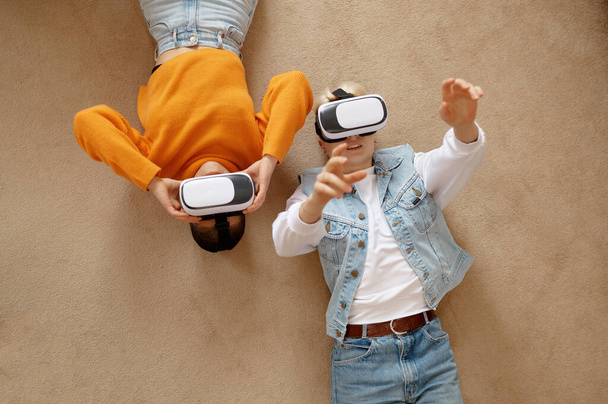 Overhead view on young attractive lesbian woman couple using vr goggles headset lying on floor at home living room. Happy girlfriends watching movie, playing games in virtual reality - Foto, afbeelding
