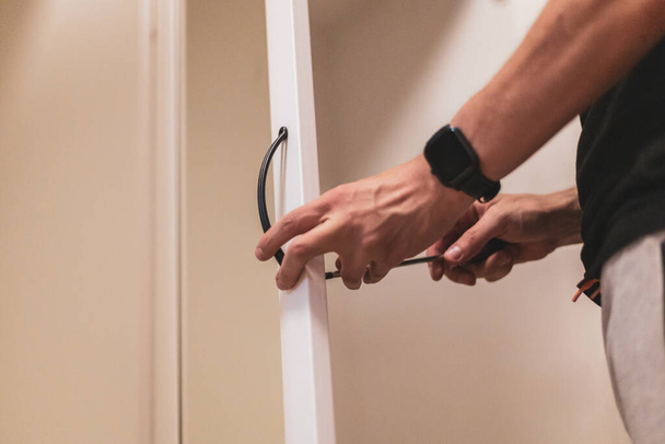 Young caucasian unrecognizable man manually installs black handles on a white wardrobe door with a mirror, tightening a screw with a screwdriver, close-up bottom view with selective focus. Furniture assembly concept. - Photo, Image