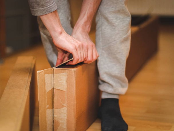 The hands of a caucasian young man in a gray tank top with sleeves open a large cardboard box in his room, close-up side view. Concept of parcel delivery, furniture assembly, shopping, at home. - Photo, Image