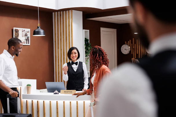 Receptionist talking to hotel guests at front desk counter in lobby, giving advice about luxury room service. Asian employee welcoming tourists at modern resort, doing check in process. - Фото, зображення