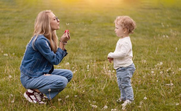 Молодая мать играет со своим маленьким сыном в парке. Одуванчики собирают цветы и дуют - Фото, изображение