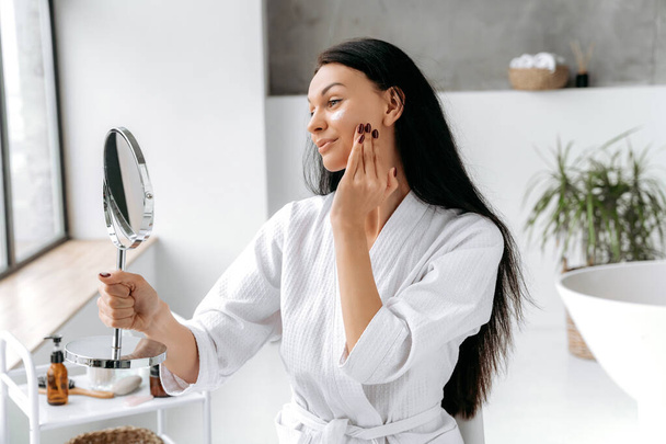 Pretty, well-groomed caucasian woman in a bathrobe, sits in a bathroom, holds a mirror, applies a moisturizing anti-aging cream on her face, prevents skin aging. Daily skin moisturizing treatments - Fotoğraf, Görsel