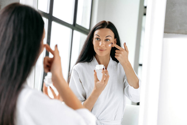 Daily skin moisturizing treatments. Beautiful well-groomed caucasian woman in a bathrobe, stands at home in front of a mirror, applies a moisturizing anti-aging cream on her face, prevents skin aging - Foto, immagini