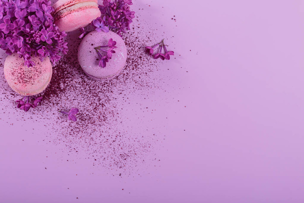 Pastel colored sweet french macaroons with lilac flowers and splash of dry blueberry powder on pink background. Beautiful composition for bakery and pastry shop. Top view with copy space - Photo, image