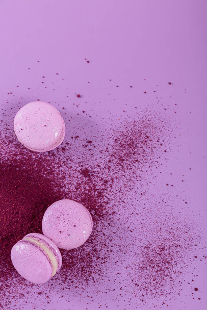Pastel colored sweet french macaroons and splash of dry blueberry powder on purple background. Beautiful composition for bakery and pastry shop. Top view with copy space for text - Photo, image