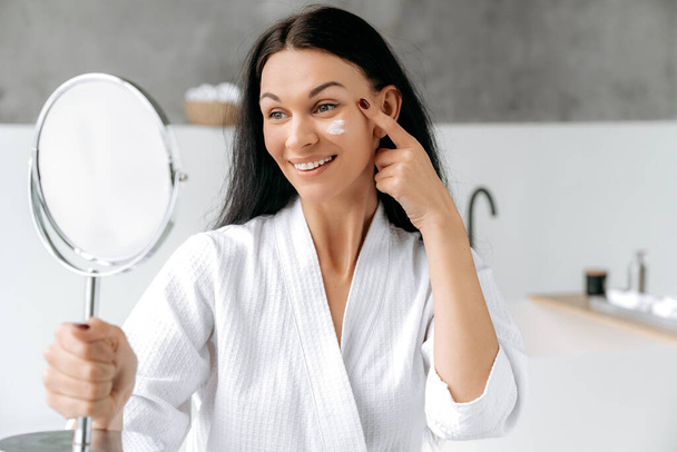 Upea hyvin hoidettu valkoihoinen nainen kylpytakissa, pitää ja katsoo peiliä, kosteuttaa anti-aging-kermaa kasvoillaan, estää ihon vanhenemista ja kuivumista. Päivittäinen iho kosteuttava hoitoja - Valokuva, kuva