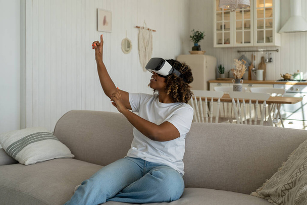 Чернокожая женщина в гарнитуре ощупывает воздух во время опыта виртуальной реальности в гостиной. Любознательная афроамериканка в шлеме, играющая дома в 3D-видеоигры, сидя на диване. Метаверсная концепция - Фото, изображение