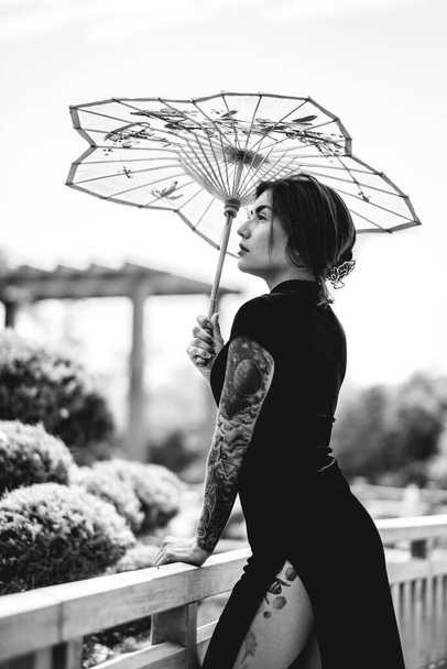 Bezaubernde Schönheit: Brünette mit Tattoos, schickem Kleid und Papierschirm, die sich an einem sonnigen Tag in einem japanischen Garten (in schwarz-weiß) erfreut) - Foto, Bild