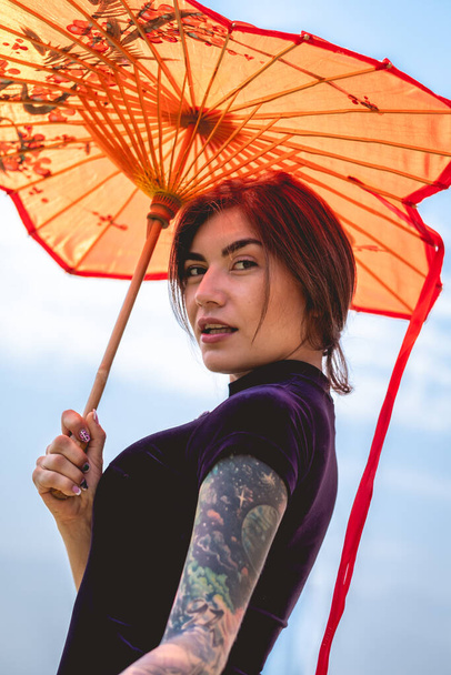 Stunning beauty: brunette with tattoos, chic dress, and red paper umbrella, delighting in a sunny day with blue sky - Photo, Image