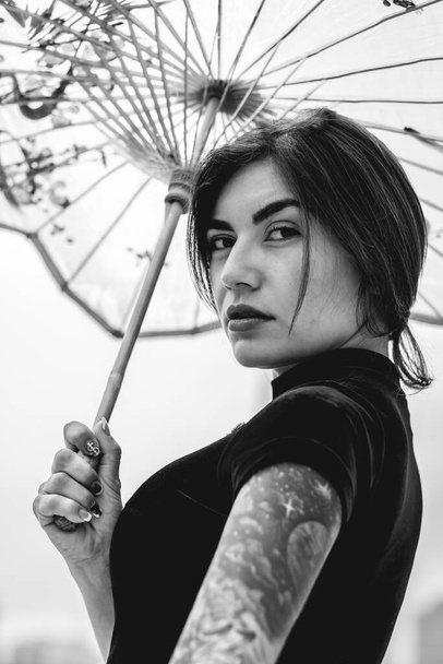 Stunning beauty: brunette with tattoos, chic dress, and paper umbrella, delighting in a pale day (in black and white) - Photo, Image