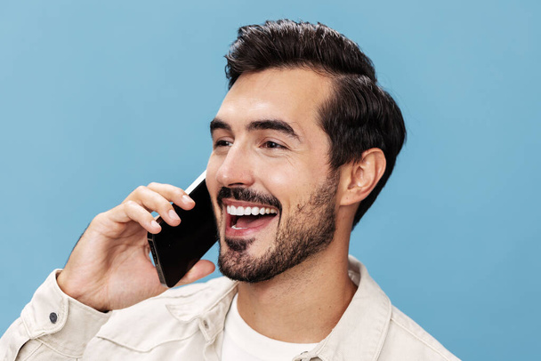 Zblízka portrét bruneta muž mluví do telefonu s kamarádem usmívá zuby, na modrém pozadí v bílém tričku a džínách, kopírovací prostor. Kvalitní fotografie - Fotografie, Obrázek