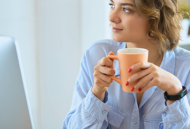 Attractive woman sitting at office desk, holding a cup of coffee. - 写真・画像