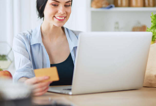 Smiling woman online shopping using computer and credit card in kitchen. - 写真・画像