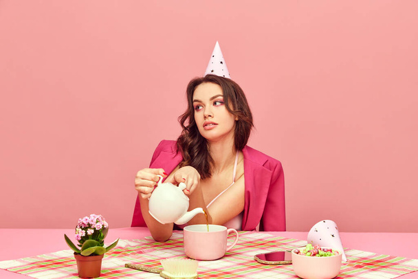 Portrait of young, beautiful, birthday girl drinking tea and eating cake against pink background. Cozy party. Concept of party, celebration, emotions, female beauty, youth. Pop art - 写真・画像
