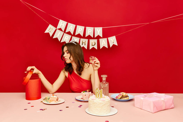 Young girl sitting at table, drinking alcohol, looking at presents and celebrating her birthday against red background. Concept of party, celebration, emotions, female beauty, youth. Pop art - Photo, image