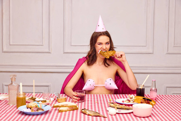Portrait of beautiful young girl with bright makeup, sitting at table, celebrating her birthday, eating chicken and drinking. Concept of party, celebration, emotions, female beauty, youth. Pop art - Photo, Image