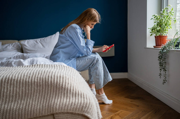 Depressed female holds cellphone pondering bad offer worried think about problem difficult solution trouble. Frustrated woman sits on bed feels upset reads bad news in message on smartphone at home. - Foto, Imagem