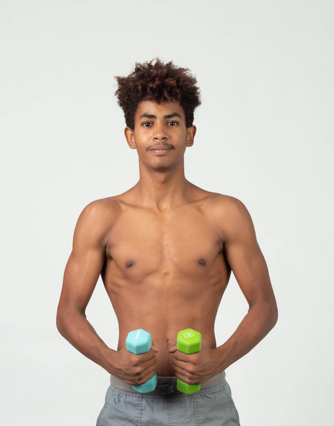 teenager s afro vlasy drží činky a ukazuje své svaly při pohledu na kameru šťastný a hrdý na svou postavu - Fotografie, Obrázek