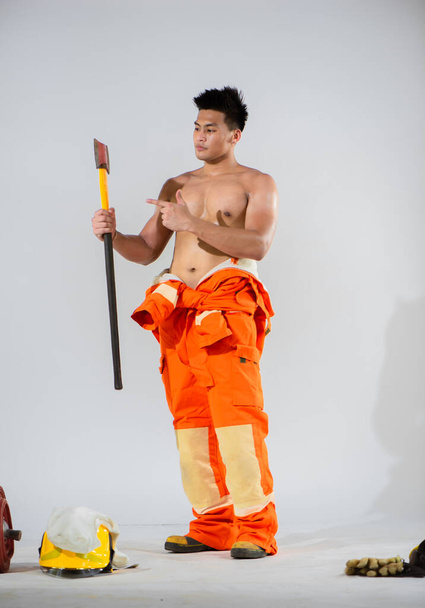 Attractive firefighter topless is standing and holding an iron axe in one hand while pointing to the axe with the other hand on a white background. - Fotó, kép
