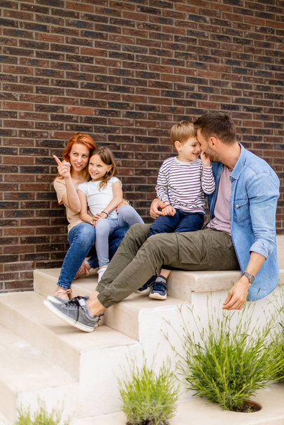 Family with a mother, father, son and daughter sitting outside on steps of a front porch of a brick house - Photo, Image