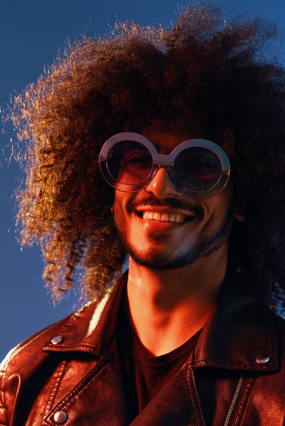 Retrato de um homem elegante com cabelo encaracolado com óculos sorrir no fundo azul multinacional, luz de cor, tendência jaqueta de couro preto, conceito moderno. Foto de alta qualidade - Foto, Imagem