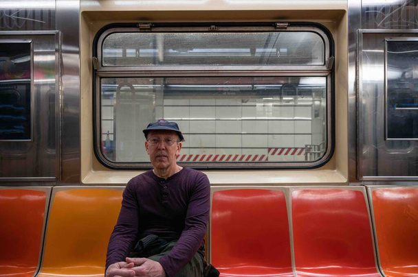 Sıradan giysili bir adam boş kırmızı koltuklarıyla tek başına metro vagonunun penceresinin önünde ve dışarıdaki metro istasyonunun önünde oturuyor.. - Fotoğraf, Görsel