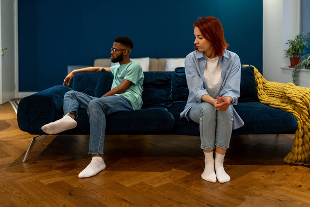 Разочарованный афроамериканец сидит на домашнем диване и смотрит на европейскую подружку после ссоры. Разнообразные пары со сложными отношениями из-за недоразумений игнорировали друг друга - Фото, изображение