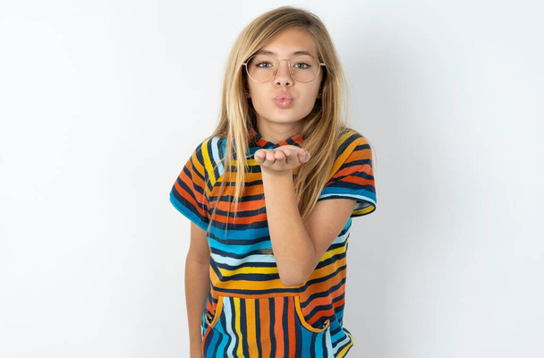 krásná teen dívka na sobě pruhované barevné tričko přes bílé pozadí studia při pohledu na kameru foukání polibek s rukou na vzduchu je krásné. Výraz lásky. - Fotografie, Obrázek