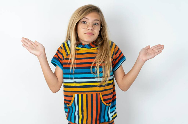 Možná ano nebo ne. Pochybné krásné teen dívka v pruhované barevné tričko přes bílé studio pozadí pokrčí rameny v údivu, snaží se učinit rozhodnutí zmatený, co chce. Váhání a nejistota. - Fotografie, Obrázek