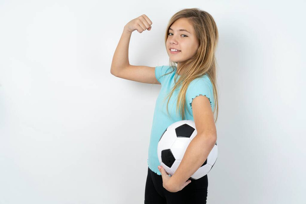 Muotokuva voimakas iloinen valkoihoinen teini tyttö yllään urheiluvaatteet tilalla jalkapallo yli valkoinen seinä osoittaa lihaksia. - Valokuva, kuva