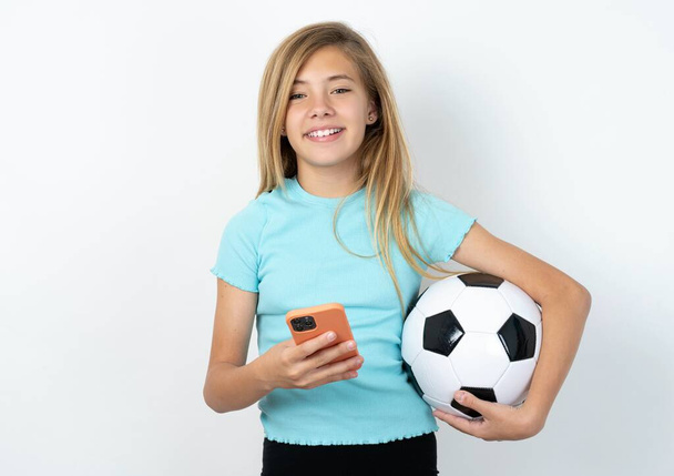 Возбужденная кавказская девочка-подросток в спортивной одежде держит футбольный мяч над белой стеной, держа смартфон и выглядя удивленной в камеру после получения хороших новостей. - Фото, изображение