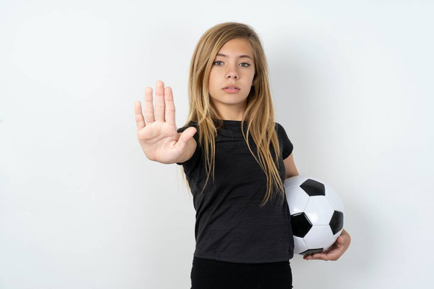 девочка-подросток в спортивной одежде держит футбольный мяч над белой стеной показывает знак стоп запрет символ держит ладонь вперед к камере со строгим выражением - Фото, изображение
