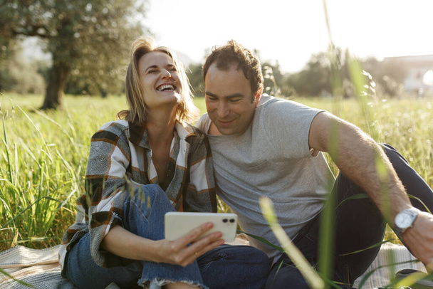 Paar, das Selfie macht oder Videoanrufe macht, während es Zeit zusammen auf einem Picknick mit Gitarre im grünen Sommergras verbringt. Partnersuche. - Foto, Bild