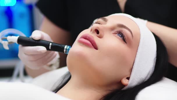 Косметологическая клиника. Профессиональная женщина-косметолог делает гидравлическую процедуру лица во время работы. Привлекательная милая женщина, лежащая на медицинской кровати во время процедур красоты - Кадры, видео