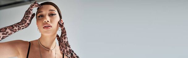 spring fashion, portrait of appealing asian woman with bold makeup, in silver necklaces, earrings and animal print gloves looking at camera on grey background, banner - Foto, Imagem