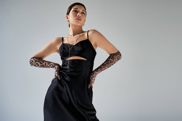 low angle view of brunette asian woman with bold makeup posing with hands on hips on grey background, black elegant strap dress, animal print gloves, silver accessories, spring fashion photography - Φωτογραφία, εικόνα