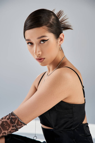 spring style asian woman with bold makeup, brunette hair and expressive gaze looking at camera on grey background, black strap dress, silver necklaces, animal print glove, fashion shoot - Φωτογραφία, εικόνα